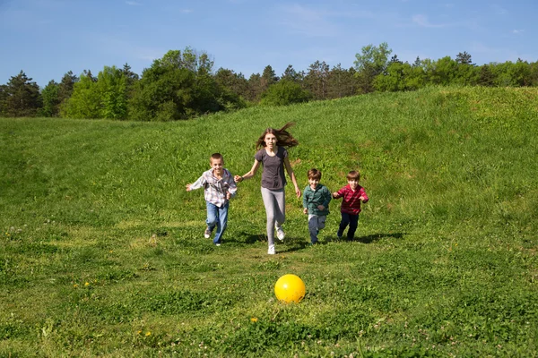 Gelukkig en lachende groep kinderen lopen naar de gele bal In het groene veld op zonnige lente — Stockfoto