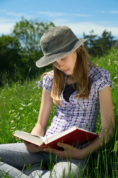 Smart tonårig flicka sitter i gräset med benen korsade läser en bok utomhus solig dag — Stockfoto