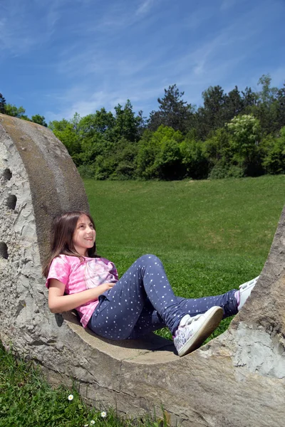 Linda chica sonriente descansando en la pared de piedra y soñando despierto en el parque en el soleado día de primavera —  Fotos de Stock