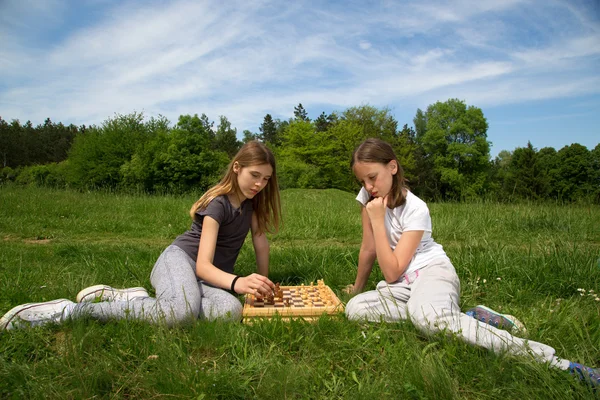 Deux filles assises sur l'herbe et jouant aux échecs, arbres forestiers et ciel nuageux bleu en arrière-plan — Photo