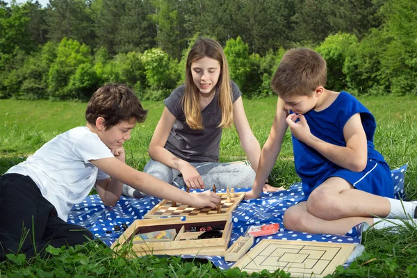 Bra sällskap sitter på gräset, pojkarna spelar schack och leende tjej njuter av spelet — Stockfoto