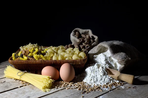 Proutěný koš naplněný těstoviny různých barev a tvarů, mouky do jutového pytle a dvě vejce na rustikální dřevěný stůl na černém pozadí — Stock fotografie