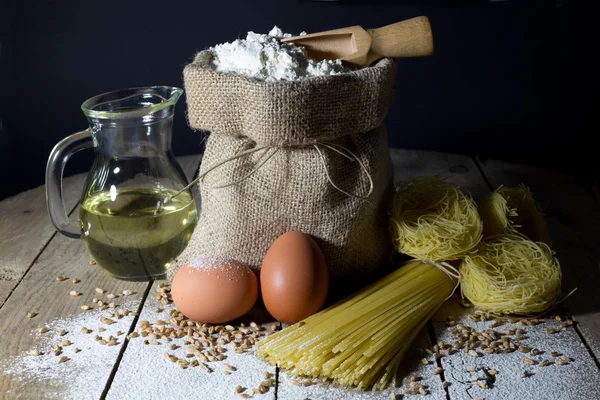 Nudeln, zwei Eier, Jutesack gefüllt mit Mehl, Kochlöffel und Olivenöl in Glasflasche auf einem alten Holztisch — Stockfoto