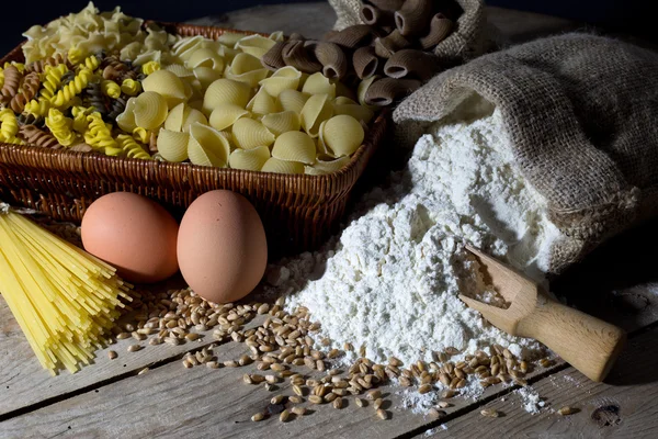 Weidenkorb gefüllt mit Nudeln in verschiedenen Farben und Formen, Mehl im Jutebeutel und zwei Eiern auf rustikalem Holztisch — Stockfoto