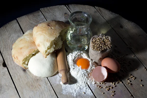 Přísady pro výrobu-mouka, olivový olej, vejce s váleček a jutového pytle naplněné pšenice na rustikální dřevěný stůl na černém pozadí — Stock fotografie