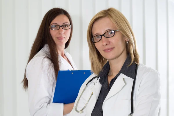 Femme médecin avec lunettes et son collègue médical avec presse-papiers en arrière-plan souriant et regardant la caméra — Photo