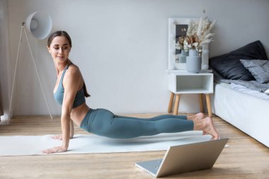 Sağlıklı kadın yoga tahtası yapıyor ve dizüstü bilgisayarda online dersler izliyor, evde oturma odasında eğitim görüyor..