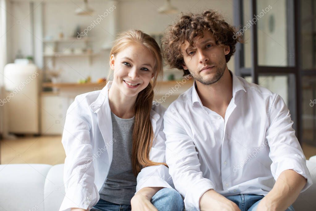 Millennial couple on couch in living room makes video call looks at camera. Distant video call.