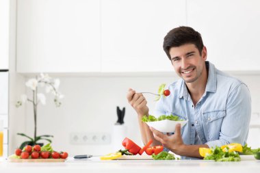 Yakışıklı adam mutfakta taze sebzelerle salata yiyor. Sağlıklı ve vejetaryen gıda konsepti.