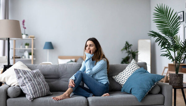 Upset young woman thinking about problem, sitting alone on couch at home, psychological problem.