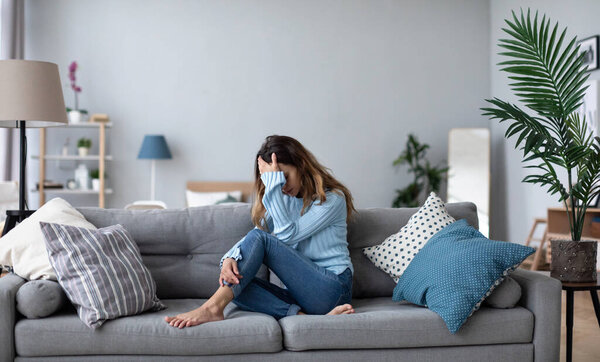Sad thoughtful worried young woman sit on sofa thinking of psychological problem. 