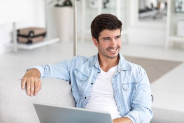 Portrait of an attractive smiling young man wearing casual clothes sitting on a couch at the living room, using laptop. Enjoying time at home. 