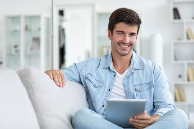 Portrait of an attractive smiling young man wearing casual clothes sitting on a couch at the living room, using digital tablet. Enjoying time at home.