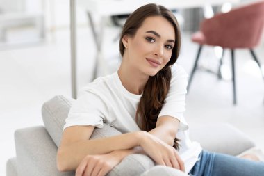 Güzel, gülümseyen otantik kadın kameraya bak, oturma odasında koltukta oturuyor, ev konforu alanı..