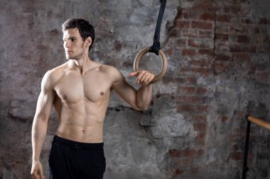Jimnastik yüzükleriyle eski tuğla duvarlı arka planda poz veren yakışıklı bir vücut geliştirici. Stüdyo çekimi.