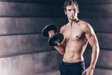 Koyu arkaplanda duran dambılları olan güzel, genç ve güçlü bir adam. Stüdyo çekimi