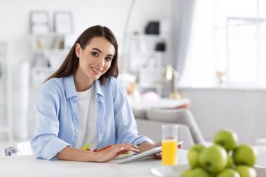 Gülümseyen kadın kahvaltıda taze sebze salatası yiyor ve evdeki mutfak masasında portakal suyu içiyor. Dijital tablette iyi haberler okuyor. Kameraya bak..