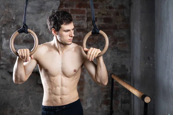 Eski tuğla duvarlı arka planda duran jimnastik yüzükleriyle yakışıklı, güçlü bir erkek fitness modeli. Stüdyo çekimi.