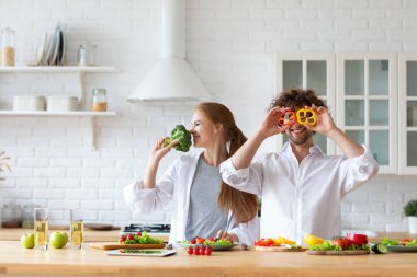 Mutfakta gülümseyen mutlu bir çift yemek pişiriyor. Eğlenceli bir adam gözlerini brokoli tutan kırmızı biberli bir kadınla kapatıyor. Sağlıklı yemek..