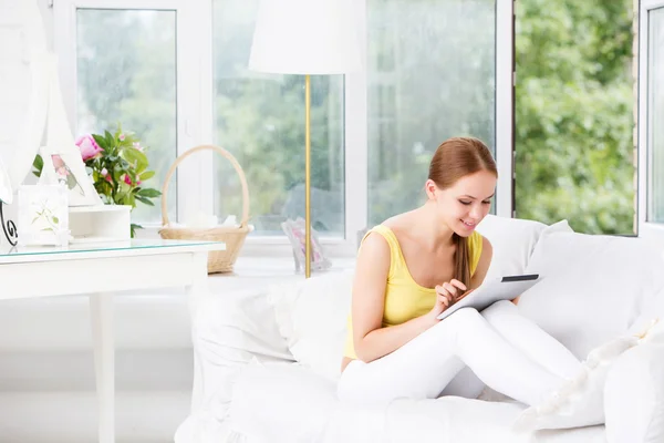 Beautiful girl sitting on a sofa against the window and shows the electronic tablet — Stock Photo, Image