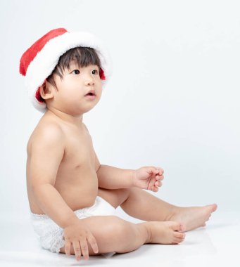 Baby Boy Noel şapkası takmış, beyaz arka planda izole bir şekilde yerde oturuyor.