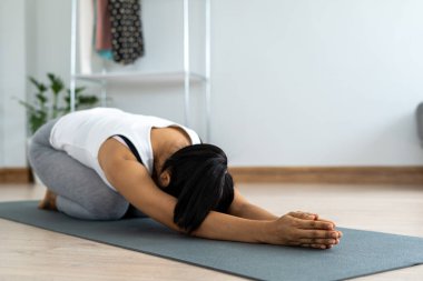 Egzersiz kıyafetli genç bir kadın evde yoga yapıyor. Kadın yerde oturuyor, kollarını açmış öne doğru eğiliyor..