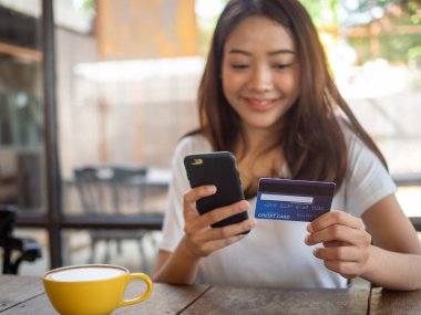 Gülümseyen genç Asyalı kadın akıllı telefon aracılığıyla online alışveriş yapmayı ve kredi kartıyla ödeme yapmayı seviyor. Nakitsiz harcama olanağı. Dikkatli olun, evden ve sosyal mesafeden alışveriş yapın.