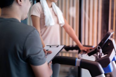 Kadın antrenör tavsiyesiyle koşu bandında egzersiz yapıyor ve aktivite detaylarını kaydediyor. Spor yaparken kaslarınızdaki yaralanma riskini azaltmak için düzgün egzersiz yapmanız tavsiye edilir..