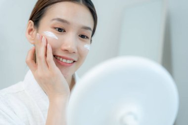 Güzel bir Asyalı kadın aynanın önünde oturur. Krem sürerek makyaj yapan sağlıklı bir kadın yüzü. Cilt kremi reklamı, kırışıklık önleyici, bebek yüzü, beyazlatma, nemlendirici, gözenekleri sıkma seru