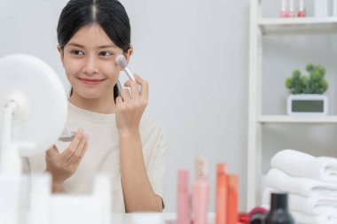 Asyalı kadın aynanın karşısına oturur ve makyaja gülümser. Makyaj yapan sağlıklı bir kadının yüzü. Reklam, yaşam tarzı, kozmetik, makyaj malzemeleri, güzellik aktiviteleri, güzellik