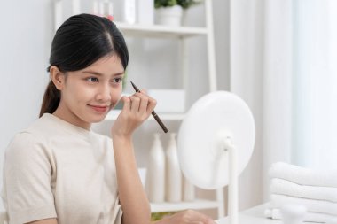 Güzel bir Asyalı kadın aynanın karşısına oturur ve makyaja gülümser. Makyaj yapan sağlıklı bir kadının yüzü. Reklam, yaşam tarzı, kozmetik, makyaj malzemeleri, güzellik etkinliği, güzellik uzmanı.