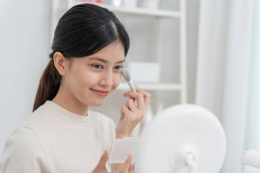 Genç Asyalı kadın aynanın karşısına oturur ve makyaja gülümser. Makyaj yapan sağlıklı bir kadının yüzü. Reklam, yaşam tarzı, kozmetik, makyaj malzemeleri, güzellik etkinliği, güzellik uzmanı.