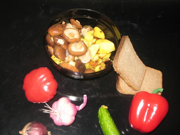 Fried potatoes, marinated mushrooms, black bread, pepper, tomato, onion, garlic, cucumber on a black background — Stock Photo, Image