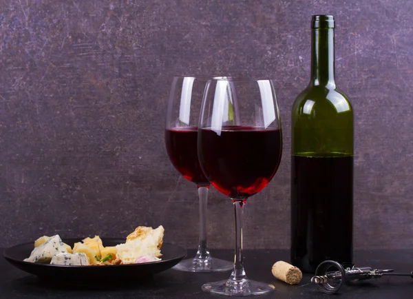 Glass and bottle of red wine, cheese, bread, garlic, nuts, salami on gray stone texture background — Stock Photo, Image