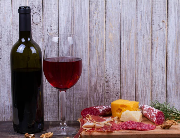 Glass and bottle of red wine, cheese, salami, walnuts, prosciutto and rosemary on wooden background — Stock Photo, Image