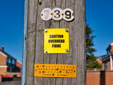 Tahta telgraf direğine sabitlenmiş sarı bir tabela optik fiber hatların tepede olduğu konusunda uyarıda bulunuyor. İngiltere 'nin kuzeybatısında güneşli bir günde çekilmiş..