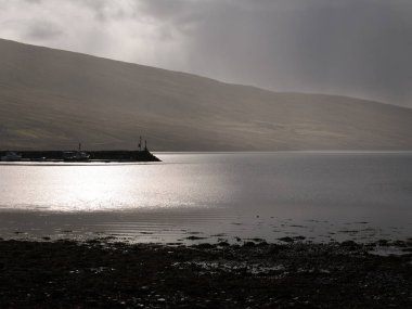 Suyun üzerindeki güneş ışığının karamsar, karamsar, karamsar, neredeyse siyah beyaz görüntüsü ve İngiltere 'nin Shetland kentindeki Olna Firth' ün başındaki iskele.