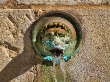 Dax, Fransa - 12 Ekim 2025: Fransa 'nın Dax kentindeki Source de la Nehe' de yıpranmış bronz bir aslan başlığından sıcak maden suyu dökülüyor. Sıcak öğleden sonra ışığı dokuyu ve ezmeyi ortaya çıkarır..
