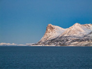 Norveç kıyılarında kışın karla kaplı dağlar açık gökyüzü altında açık mavi bir denizin üzerinde yükseliyor, temiz ve minimalist bir ruh hali içinde..