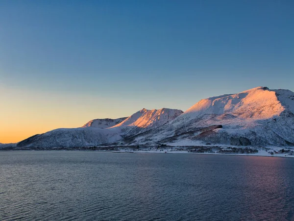Altın saat ışığı karla kaplı dağları ısıtıyor Norveç kıyısındaki sakin bir kıyı şeridinin üzerinde, kışın kışın kışın serin göğü ve soğuk mavi suyla.