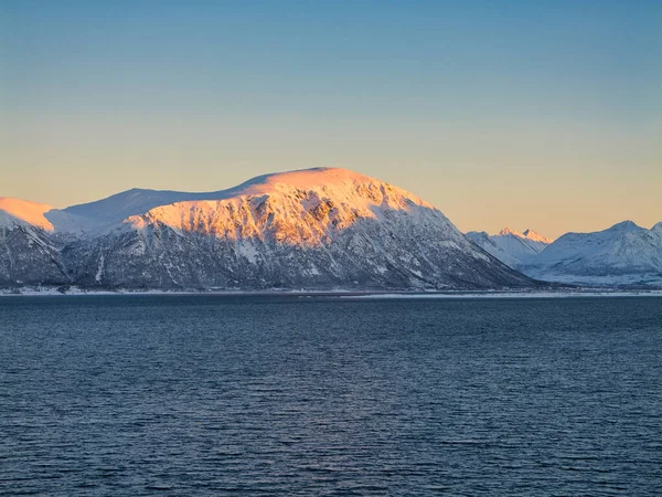 Karlı dağlar, Norveç kıyısındaki karanlık bir fiyordun üzerinde, yumuşak kış güneşinde parlayarak sakin, minimalist bir İskandinav manzarası yaratıyor..
