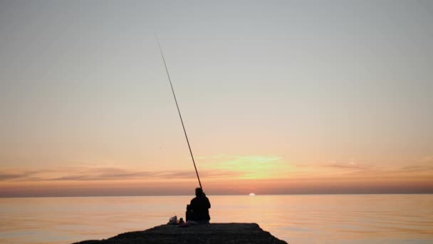 夕暮れ時の漁師のシルエットは海の桟橋に座って釣竿で魚を捕る 明るいオレンジ色の夜明けは湖の上の澄んだ夏の空を照らし — ストック動画