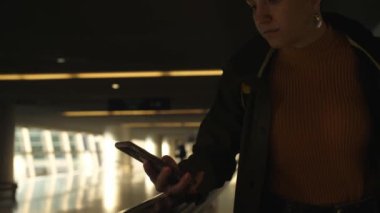 Sarı ceketli ve sabah yeşil yağmurluklu genç bir kadın havaalanındaki yürüyen merdivende duruyor ve akıllı telefon kullanıyor ve gülümsüyor. Şafak yüzünü aydınlatıyor