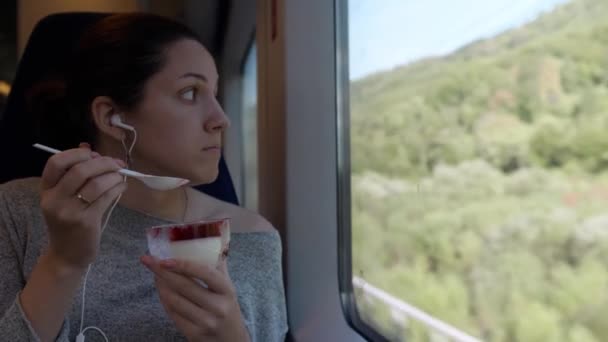 黒い髪の少女が窓の近くの電車に座ってデザートを食べています 窓の外には緑の夏の森が広がっています — ストック動画