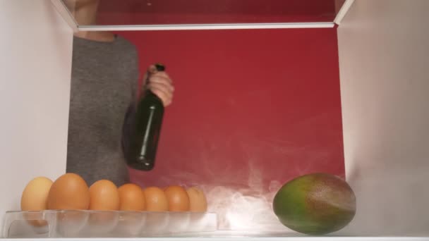 白い女が冷蔵庫に入り冷蔵庫の棚に緑色のビールを入れてる 冷蔵庫には煙が充満していて棚の上の新鮮な果物の上にゆっくりと広がり — ストック動画