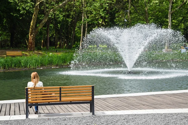 Young girl is resting on a park bench next to a fountain — Stock Photo, Image