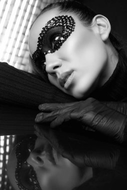 Siyah balıkçı yaka kazaklı ve pullu maskeli zarif esmer kadının portresi stüdyoda aynalı bir yüzeye poz veriyor.