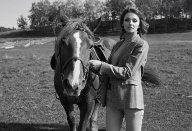 Zarif, kahverengi ceketli, kır manzarasında bir atla poz veren esmer bir kadının portresi.