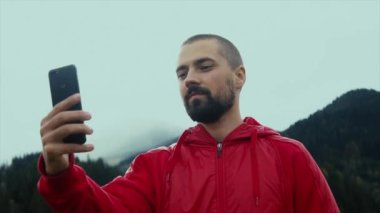 Elinde telefonla dağların arka planına bakan sakallı, beyaz bir adam. Gezgin görüntülü arama. Blogcu doğada yayın yapıyor..