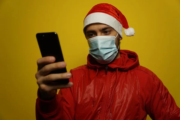 Elinde telefon olan modern bir Noel Baba 'nın portresi. Sarı arka planda izole edin. Tıbbi maske.